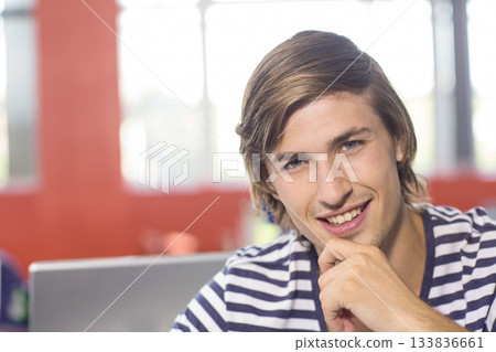 Male student smiling at camera, resting chin on hand in classroom with silver laptop, copy space 133836661
