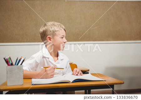 Elementary school boy sitting at wooden desk in classroom using open workbook and colored pencils Elementary school boy sitting at wooden desk in classroom using open workbook and colored pencils 133836670