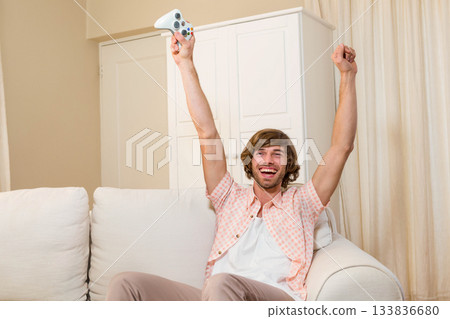 Adult man raising arms triumphantly sitting on white sofa in living room with white game controller Adult man raising arms triumphantly sitting on white sofa in living room with white game controller 133836680
