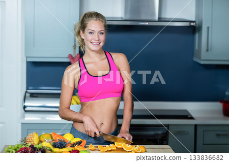 Woman wearing pink sports bra and gray leggings chopping oranges on wooden board in modern kitchen 133836682