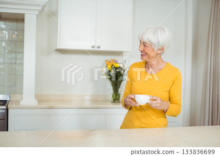 Senior woman standing behind marble countertop holding white mug in home kitchen, copy space 133836699