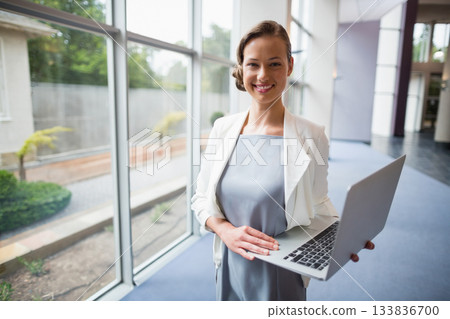Businesswoman standing in sleek office corridor beside glass windows, holding silver laptop Businesswoman standing in sleek office corridor beside glass windows, holding silver laptop 133836700