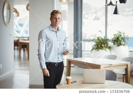 Man wearing business attire reviewing files on tablet in office lounge with laptop coffee cup 133836706