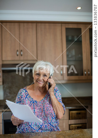 Senior woman holding documents and talking on smartphone in kitchen with polished stone countertop 133836707