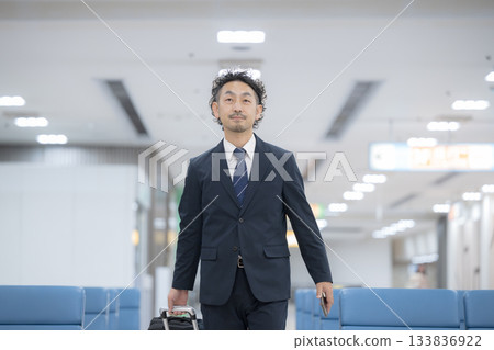 A middle-aged businessman in a suit carrying a suitcase or carry-on bag walking through an airport or station 133836922