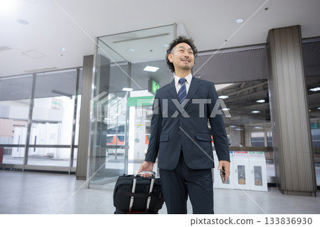 A middle-aged businessman in a suit carrying a suitcase or carry-on bag walking through an airport or station 133836930