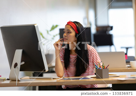 Asian woman wearing headset and striped shirt sitting at modern office desk typing on keyboard Asian woman wearing headset and striped shirt sitting at modern office desk typing on keyboard 133836971