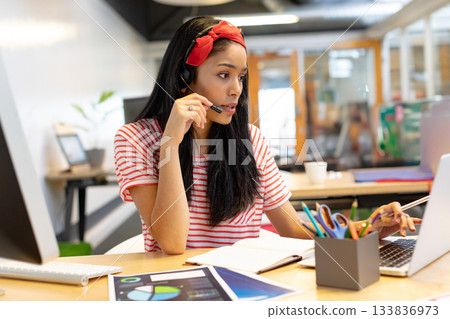African American woman typing on laptop at office desk wearing headset and reviewing printed charts 133836973