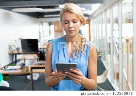 Woman wearing sleeveless blue blouse standing and holding tablet computer in office near desks Woman wearing sleeveless blue blouse standing and holding tablet computer in office near desks 133836978