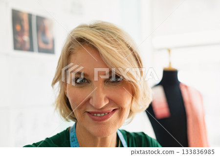 Middle-aged woman smiling and wearing tape measure in studio with dress form and pinned sketches 133836985