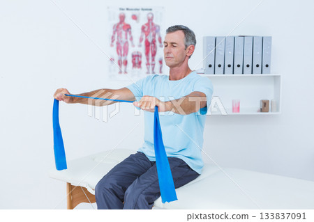 Middle-aged man pulling blue elastic band on table in therapy room with skeletal chart, shelf, cup Middle-aged man pulling blue elastic band on table in therapy room with skeletal chart, shelf, cup 133837091