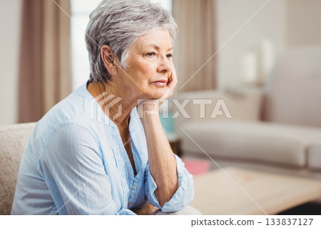 Senior woman sitting on sofa supporting cheek, gazing at coffee table at home, copy space Senior woman sitting on sofa supporting cheek, gazing at coffee table at home, copy space 133837127