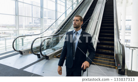 Male business traveler standing gripping suitcase in airport terminal near escalators, copy space Male business traveler standing gripping suitcase in airport terminal near escalators, copy space 133837313