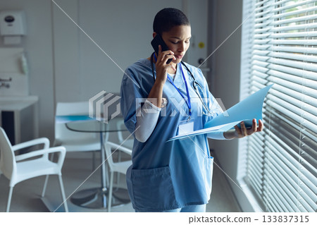 African American female nurse speaking on smartphone holding folder wearing stethoscope in clinic 133837315