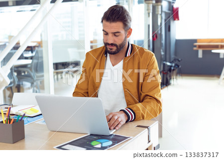 Male wearing bomber jacket working at shared office desk using laptop and viewing printed charts Male wearing bomber jacket working at shared office desk using laptop and viewing printed charts 133837317