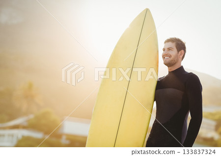 Male surfer standing on shoreline at sunset holding yellow surfboard in black wetsuit, copy space Male surfer standing on shoreline at sunset holding yellow surfboard in black wetsuit, copy space 133837324