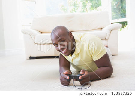 African American man lying at home using black wired controller smiling by window with foliage 133837329