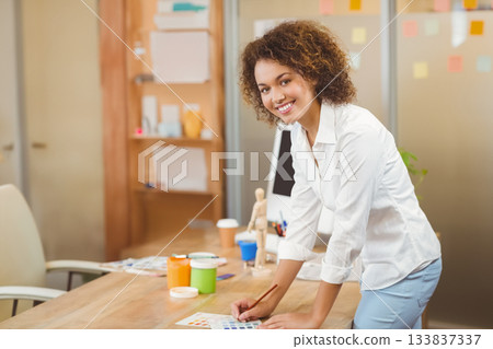 African American woman leaning over desk reviewing swatches with brush and paint jars in studio African American woman leaning over desk reviewing swatches with brush and paint jars in studio 133837337