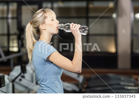 Female gym-goer drinking water from reusable bottle by treadmills in gym, copy space Female gym-goer drinking water from reusable bottle by treadmills in gym, copy space 133837345
