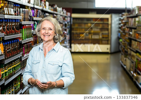 Senior woman holding smartphone and checking product info in supermarket aisle of condiment shelves Senior woman holding smartphone and checking product info in supermarket aisle of condiment shelves 133837346