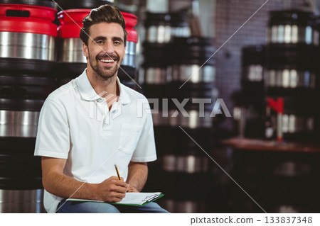 Male brewery technician sitting on stool in storage area taking clipboard notes by stainless kegs 133837348