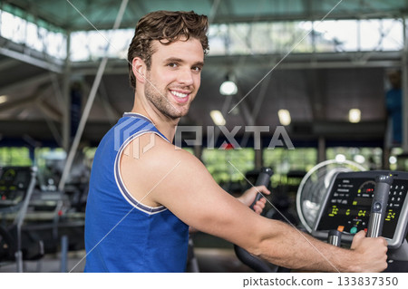 Man smiling and exercising on elliptical trainer with digital display under overhead lights in gym 133837350