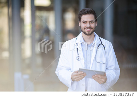 Male doctor smiling and holding tablet standing in hospital lobby wearing lab coat and stethoscope 133837357