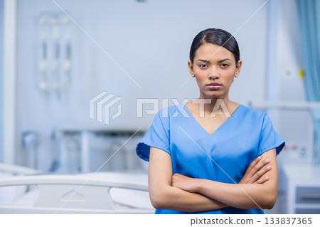 Female nurse standing confidently with arms crossed in hospital room with bed rails and IV pole 133837365