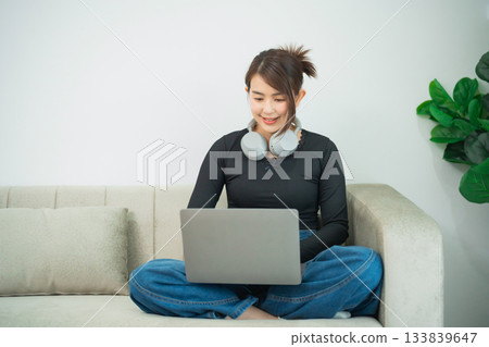 Young woman working on laptop at home with headphone on, sitting on couch in casual outfit, focused on screen, indoor workspace with plants around. Young woman working on laptop at home with headphone on, sitting on couch in casual outfit, focused on screen, indoor workspace with plants around. 133839647