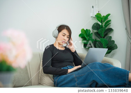 Young Woman with Headphones Using Laptop at Home, Engaged in Conversation, Relaxed Atmosphere with Indoor Plants and Soft Lighting Young Woman with Headphones Using Laptop at Home, Engaged in Conversation, Relaxed Atmosphere with Indoor Plants and Soft Lighting 133839648