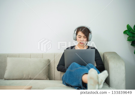 Young woman wearing headphones engaging with laptop while sitting comfortably on sofa in modern living room, showcasing work-from-home lifestyle Young woman wearing headphones engaging with laptop while sitting comfortably on sofa in modern living room, showcasing work-from-home lifestyle 133839649