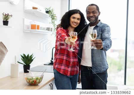 A smiling couple in a kitchen cheers with glasses of white wine, celebrating together. 133839662