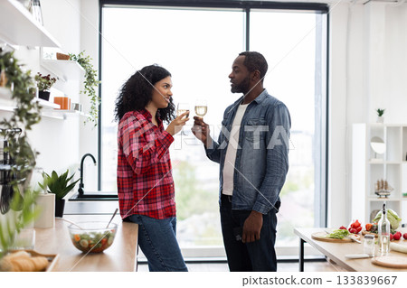 A diverse couple enjoys a moment together, toasting with wine glasses in a bright, contemporary kitchen setting. 133839667