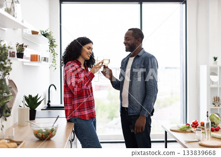 A smiling couple clinks glasses of wine in a bright, modern kitchen, celebrating a special moment together. 133839668