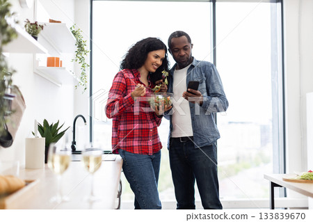 A diverse couple enjoys a healthy meal together in a modern kitchen, with one looking at a phone. 133839670