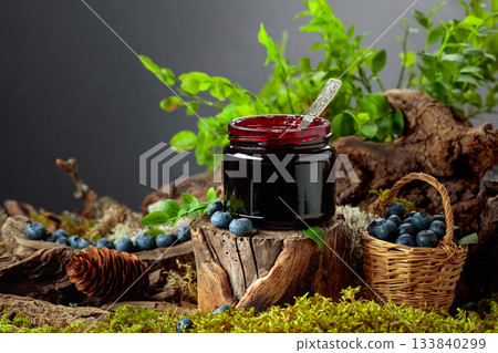 A jar of blueberry jam with fresh berries on an old tree trunk in the forest. 133840299