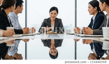 Asian businesswoman leading team meeting with diverse colleagues around reflective conference table in modern office boardroom 133840349