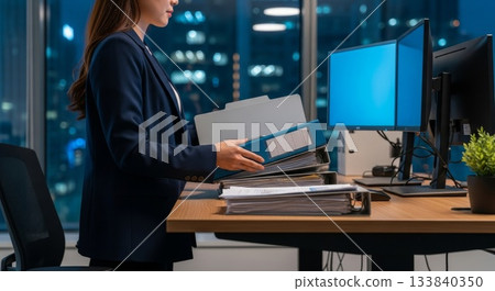 Asian businesswoman organizing financial documents and files in modern corporate office during evening work hours with city skyline view 133840350
