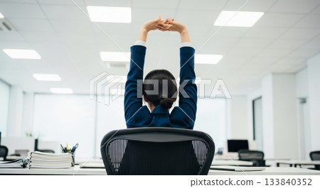 Asian businesswoman stretching arms overhead while sitting in ergonomic office chair during work break in modern corporate workplace with fluorescent lighting 133840352