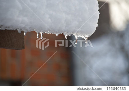 Icicles Hanging From Snow-Covered Roof Over Wooden Beam With Blurred Brick Wall Background Icicles Hanging From Snow-Covered Roof Over Wooden Beam With Blurred Brick Wall Background 133840883