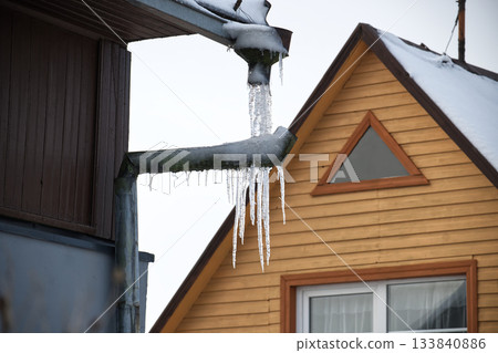 Icicles Hanging From Snowy Roof Pipes Between Dark Wooden Building And Bright Wooden Cottage Icicles Hanging From Snowy Roof Pipes Between Dark Wooden Building And Bright Wooden Cottage 133840886