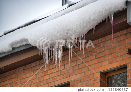 Icicles hanging from a snow-covered roof against a brick wall in winter Icicles hanging from a snow-covered roof against a brick wall in winter 133840887