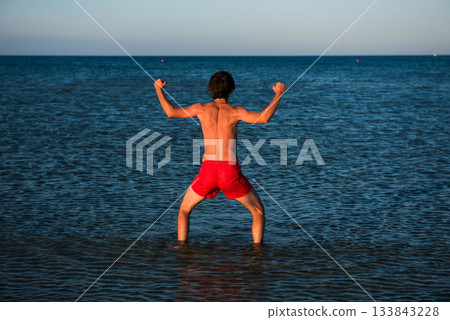 Slim guy posing in red swimwear in sea water Slim guy posing in red swimwear in sea water 133843228