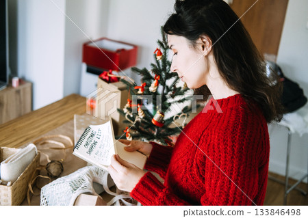 Young adult woman in red sweater reads handwritten Christmas shopping list on notebook next to small decorated tree. Holiday planning, conscious consumerism, Christmas shopping, mindful gifting. Young adult woman in red sweater reads handwritten Christmas shopping list on notebook next to small decorated tree. Holiday planning, conscious consumerism, Christmas shopping, mindful gifting. 133846498