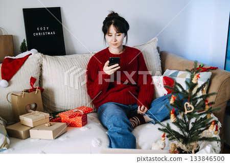Young adult woman sits on a couch using a mobile phone with Christmas gifts and a small decorated tree nearby. Mobile commerce, smartphone buying habits, holiday m-commerce, virtual shopping. Young adult woman sits on a couch using a mobile phone with Christmas gifts and a small decorated tree nearby. Mobile commerce, smartphone buying habits, holiday m-commerce, virtual shopping. 133846500