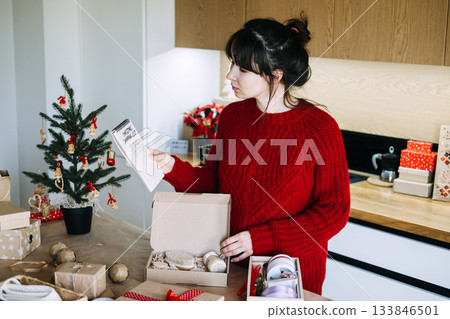 Young adult woman reads handwritten Christmas shopping list on notebook while preparing gift box on kitchen counter. Christmas shopping list, planned gifting, budgeting gifts, holiday organization. 133846501