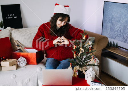 Woman in a Santa hat forms a heart gesture with her hands while seated near a small Christmas tree during a video call. Digital and physical connection, hybrid holiday traditions, remote gifting Woman in a Santa hat forms a heart gesture with her hands while seated near a small Christmas tree during a video call. Digital and physical connection, hybrid holiday traditions, remote gifting 133846505