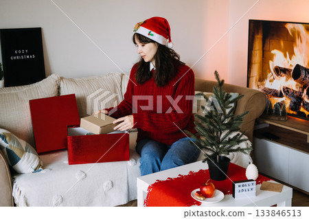 A young woman sits on a sofa decorating a small tabletop Christmas tree with handcrafted straw ornaments. Reusable decor, natural ornaments, sustainable home accents, low-impact Christmas. A young woman sits on a sofa decorating a small tabletop Christmas tree with handcrafted straw ornaments. Reusable decor, natural ornaments, sustainable home accents, low-impact Christmas. 133846513