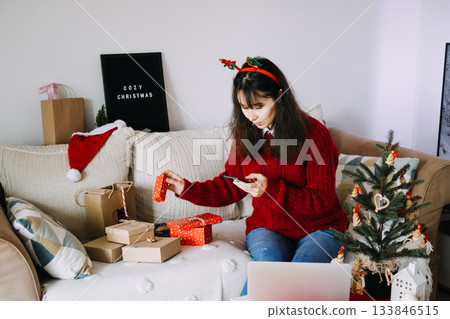 Young woman in red sweater shops for Christmas gifts on her smartphone while sitting on couch. Mobile commerce, smartphone holiday shopping, cozy Christmas, online consumer behavior Young woman in red sweater shops for Christmas gifts on her smartphone while sitting on couch. Mobile commerce, smartphone holiday shopping, cozy Christmas, online consumer behavior 133846515