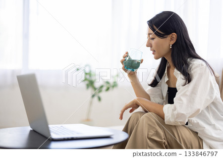 一名女子一邊喝水一邊看著筆記型電腦。 133846797